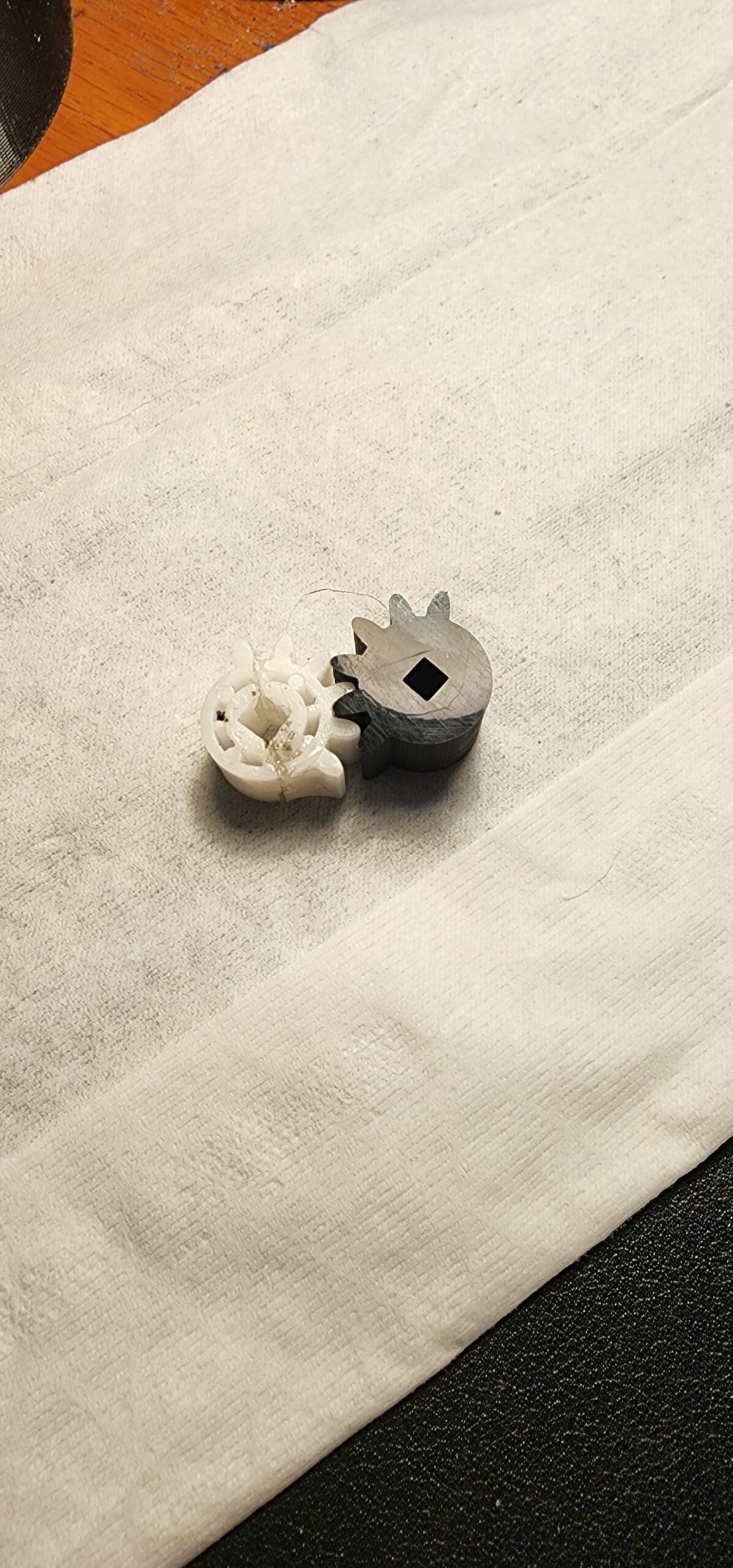 Damaged white original gear beside Kevin’s gray 3D-printed prototype with a square center opening